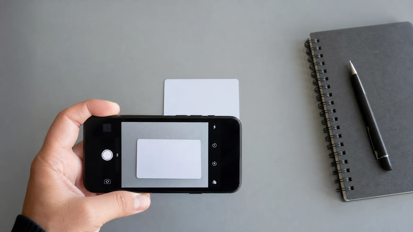 Hand photographing a blank identity card lying flat on a desk beside a notebook and pen